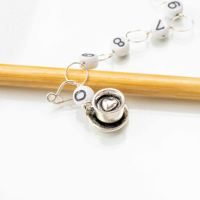 Close-up of silver tea cup with heart latte art charm on numbered bead chain row counter resting on bamboo knitting needle