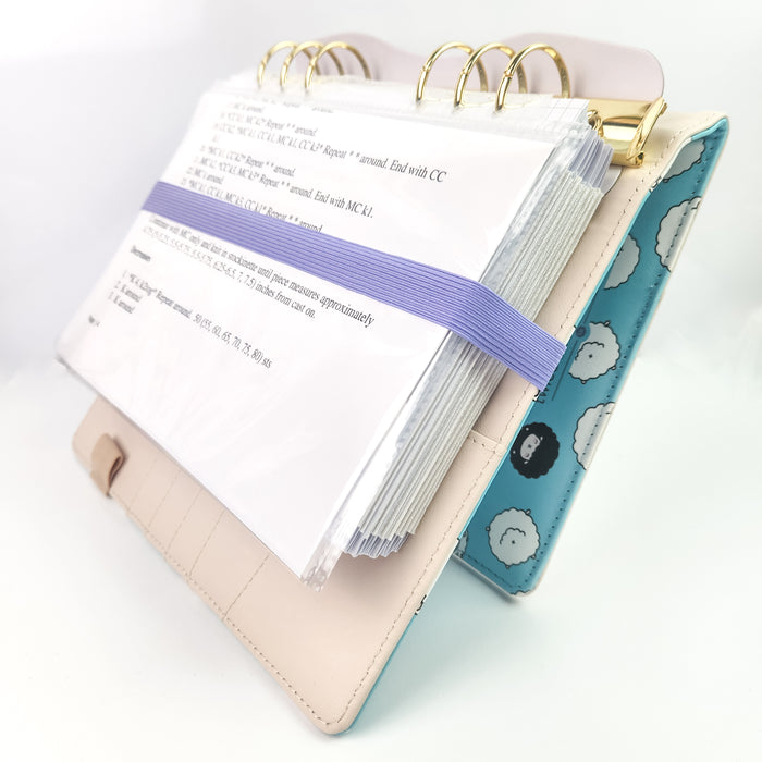 Project Journal Planner standing open tent-style showing printed knitting pattern pages in plastic sleeves with a lavender elastic band and teal sheep cover visible on the back