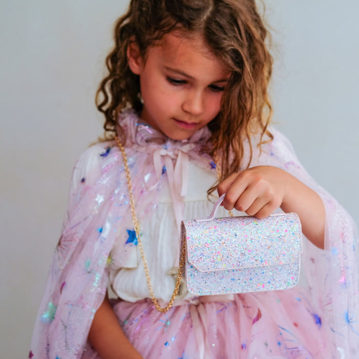 A young girl with curly brown hair looks down at a small glittery pink purse she is holding. She is wearing a pink sparkly cape and a white ruffled blouse with a bow tie.