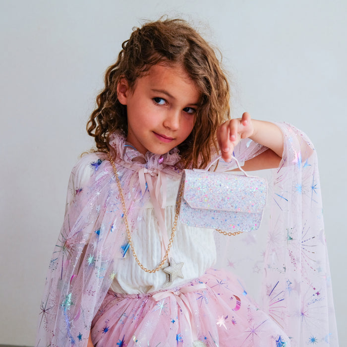 A young girl with curly brown hair is wearing a whimsical light pink and white fairy-tale style dress with sheer star-patterned sleeves, holding a matching sparkly handbag.