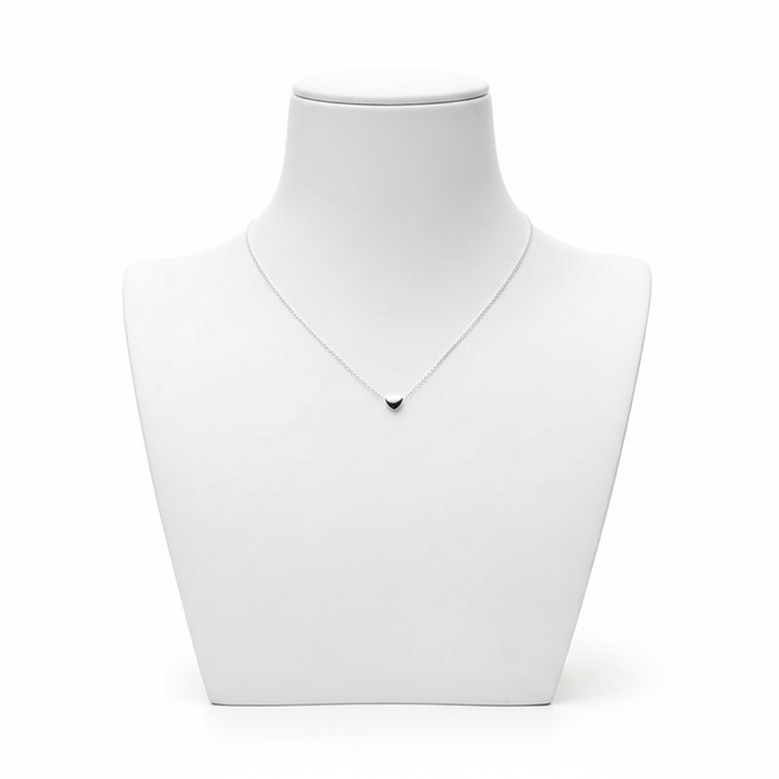 A high-resolution product shot of a minimalist silver necklace with a tiny, solid heart-shaped pendant on a fine link chain, displayed on a clean white jewelry bust against a pure white background.