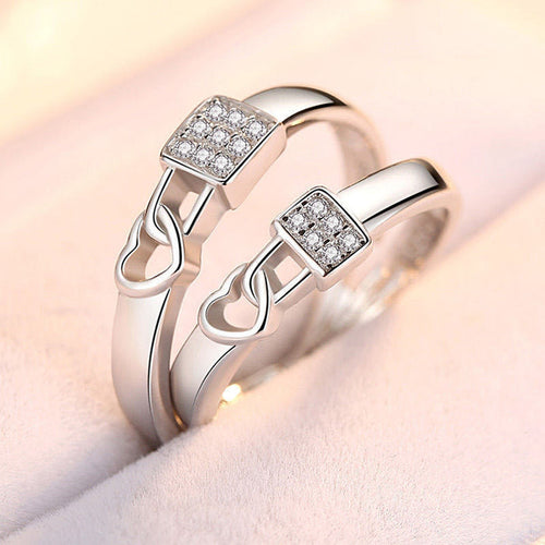 Close-up of two polished silver-tone rings for men and women, featuring a square-shaped crystal setting and a delicate dangling heart charm, displayed in a soft-lit luxury jewelry box.