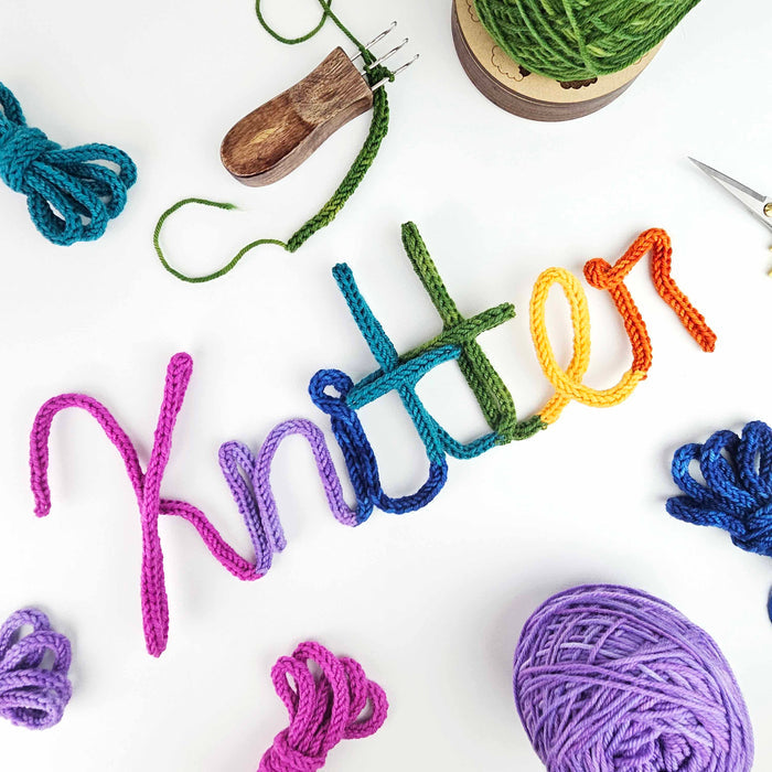 Rainbow i-cord wire art spelling Knitter in cursive with Cord Quill tool, yarn balls, and scissors