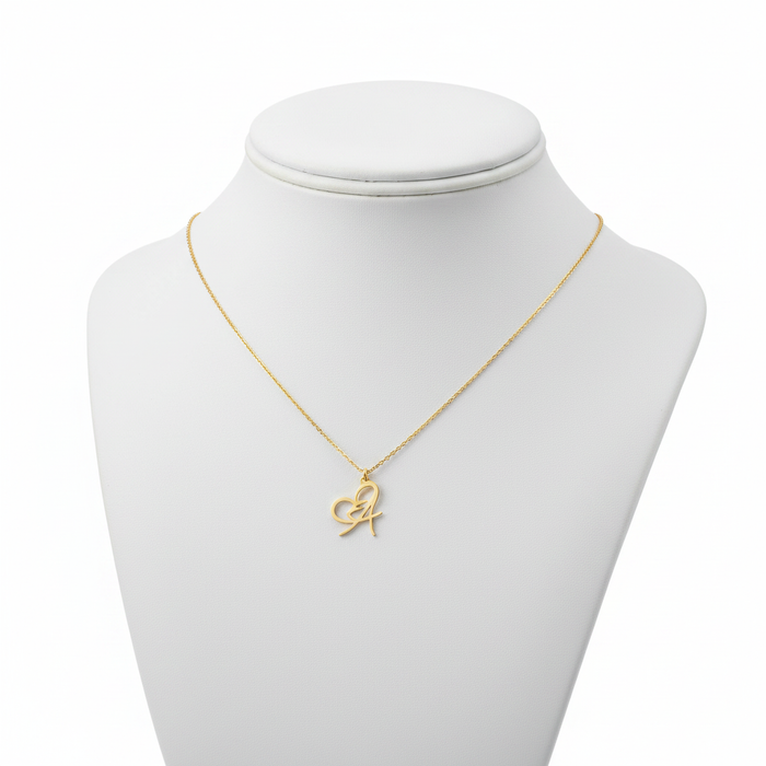 A gold-toned necklace featuring a delicate pendant that blends a script-style initial "A" with an integrated open-heart design. The pendant is suspended from a fine gold cable chain and displayed on a white jewelry mannequin bust against a pure white background.