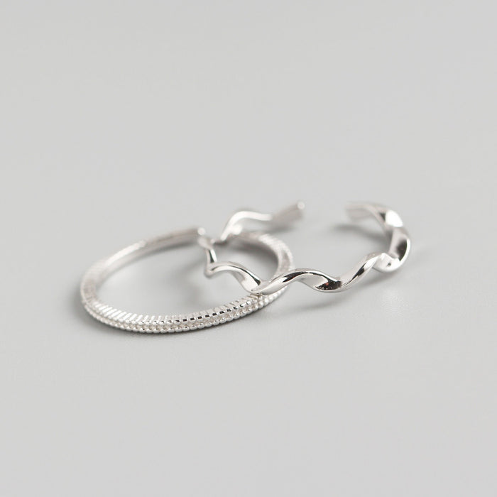 High-angle product photography of two silver-tone rings on a charcoal grey surface; one ring features a consistent beaded texture and the other a polished, undulating wave design with an open back.