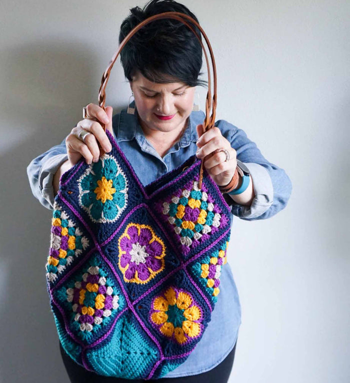 Woman holding colorful granny square crochet tote bag in teal, purple, gold, and navy with leather straps
