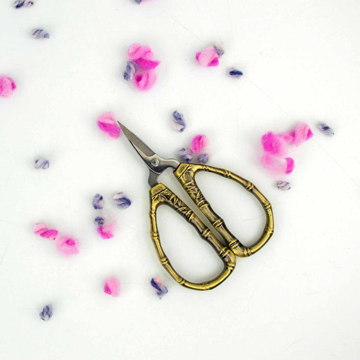 Bronze bamboo handle thread snips tiny scissors with pink and purple wool confetti on white background