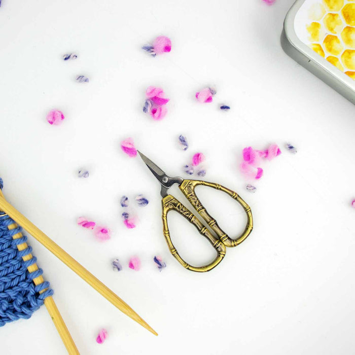 Bronze bamboo handle thread snips scissors with blue knitting project, wool confetti, and honeycomb stitch marker tin
