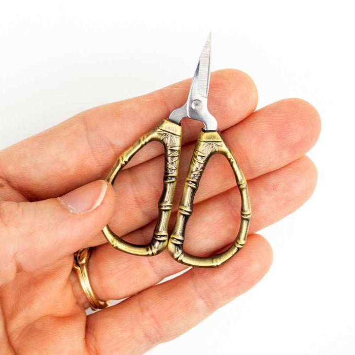 Bronze bamboo-handled thread snips held in hand showing the small ornate scissors with finger loops and sharp blades