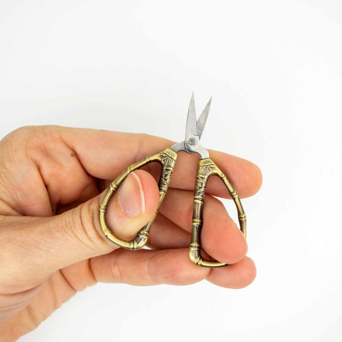 Hand holding bronze bamboo thread snips open to show the sharp scissor blades, with ornate bamboo-shaped handles