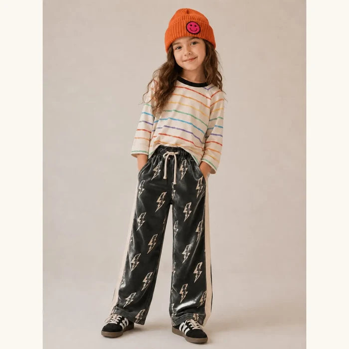 A young girl stands against a plain beige studio backdrop wearing an orange knit beanie with a pink smiley face, a cream long-sleeve striped shirt, black wide-leg pants with a white drawstring and repeated white lightning-bolt print, and black-and-white sneakers.