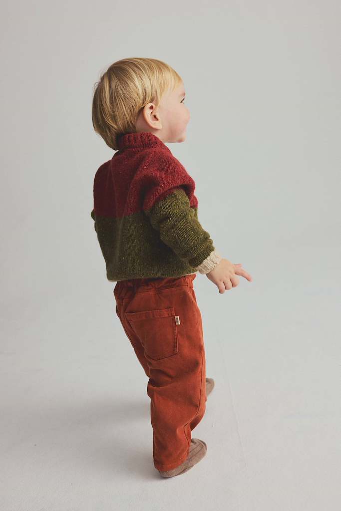 A toddler standing in profile wearing a color-blocked knit sweater with a burgundy top half, olive green bottom half, and rust-orange pants, photographed against a plain light gray studio background.