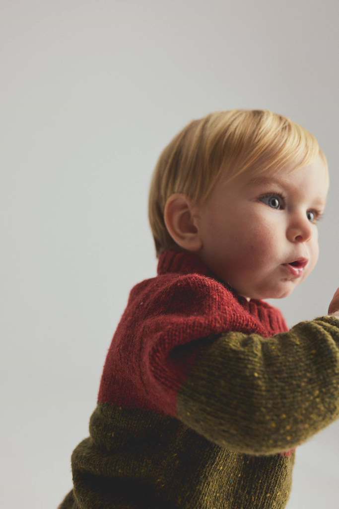 A close-up side profile of a young toddler with blonde hair wearing a color-block knit sweater with a red yoke and olive green sleeves, set against a plain light gray background.