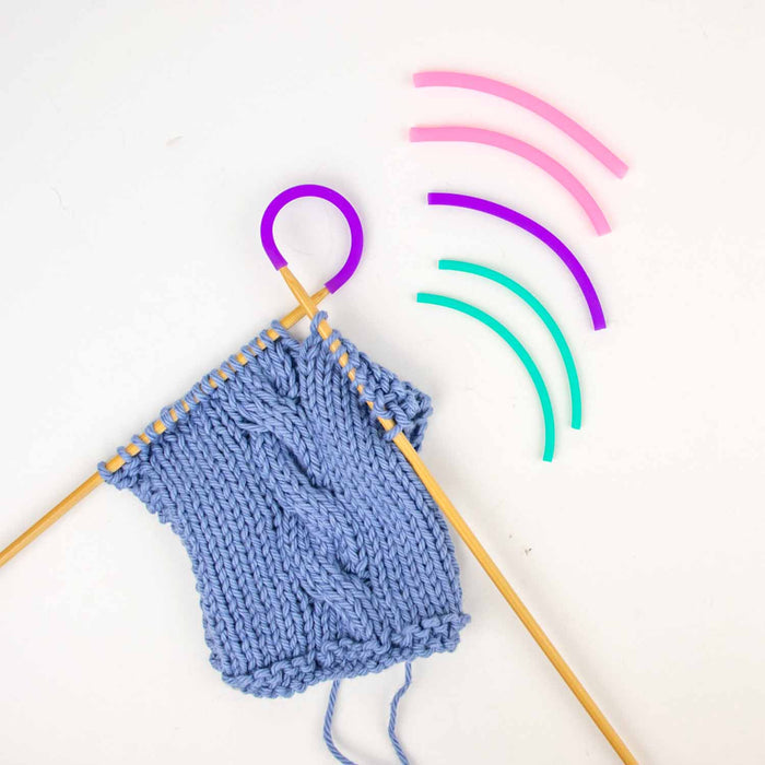 Blue cable knit swatch on bamboo needles with purple Tip Tie attached and three extra Tip Tie pairs in teal, purple, and pink beside it