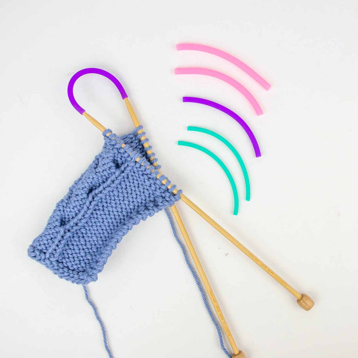 Blue cable knit swatch on bamboo needles with purple Tip Tie and three Tip Tie pairs in teal, purple, and pink shown from an alternate angle