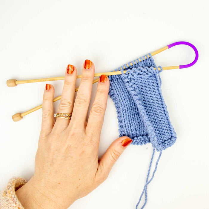 Hand holding a blue cable knit swatch on bamboo needles with purple Tip Tie point protector showing the secure hold during transport