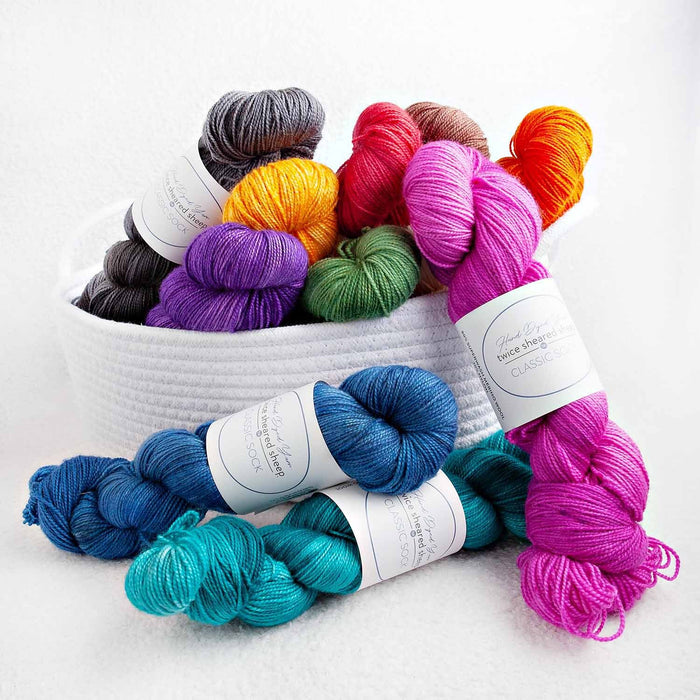 White basket overflowing with colorful skeins of Twice Sheared Sheep Classic Sock hand-dyed fingering weight yarn in blue, teal, pink, purple, orange, green, and gray