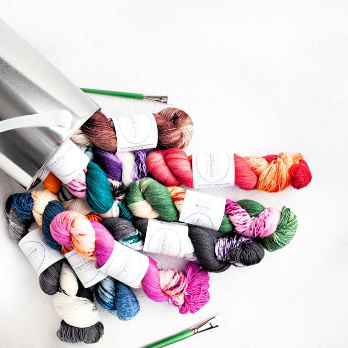Colorful collection of TSS Classic Sock Paints hand-dyed variegated yarn skeins spilling from a white tin onto a white background with green crochet hooks