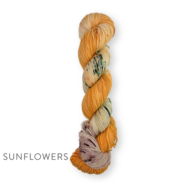 Single skein of TSS Classic Sock Paints yarn in Sunflowers colorway featuring warm golden yellow and amber sections with green speckles and soft lavender on a white background