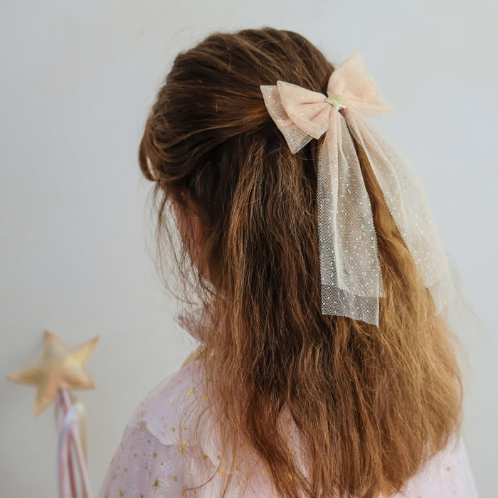 A child's long brown hair styled with a pale pink bow and sheer white tulle ribbon, shown from the back against a light gray background.