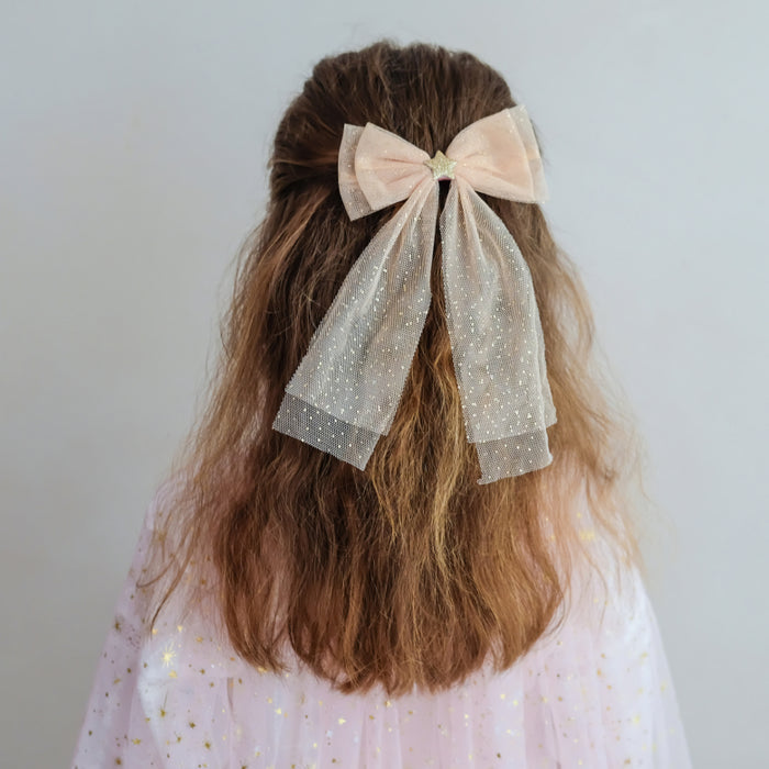 A child's long, wavy brown hair styled with a large pale pink and sheer glittery bow accessory, shown from the back against a plain light gray background.