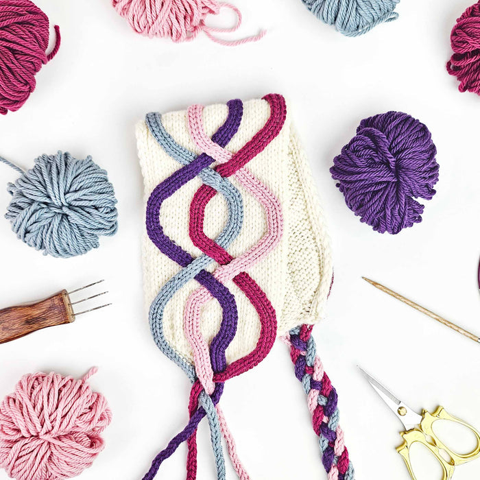 Flat lay of the Twining Roads Headband in cream with intertwining purple, pink, rose, and blue cord quill cables with yarn balls, scissors, and a cord quill tool
