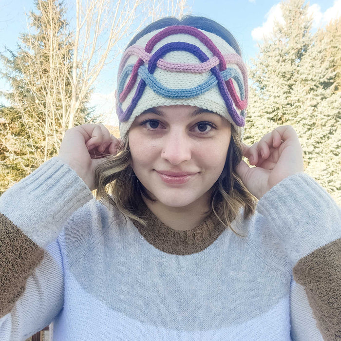 Model wearing the Twining Roads Headband in cream with colorful purple, pink, and blue intertwining cord quill cables, front view outdoors