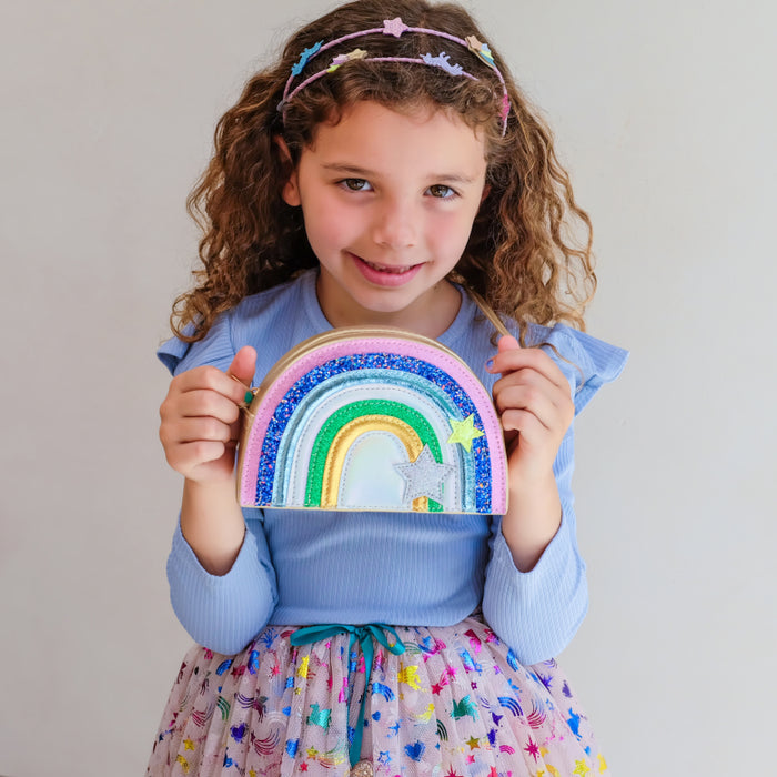 A young girl with curly brown hair wearing a pastel rainbow headband and a blue top holds a small, glittery rainbow-shaped pouch or bag against a plain light gray background.
