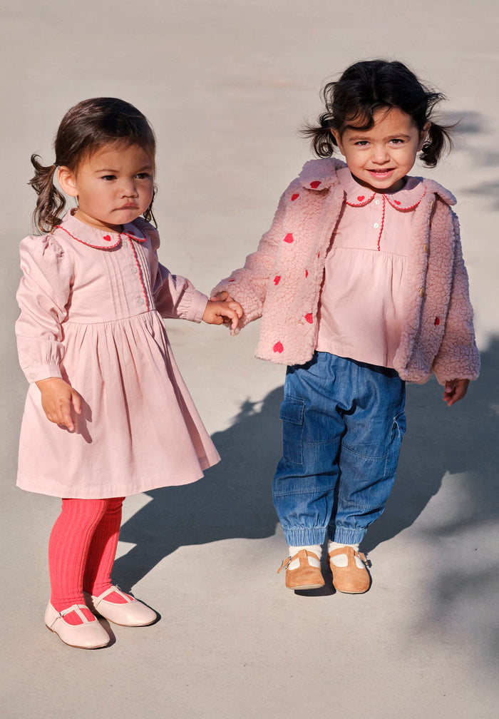 Two young children holding hands, wearing matching pink outfits with blue pants.
