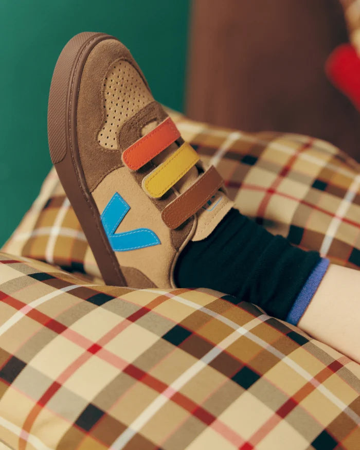 A close-up of a child's foot wearing a brown suede sneaker with colorful velcro straps and a blue V logo, resting on a beige plaid cushion.