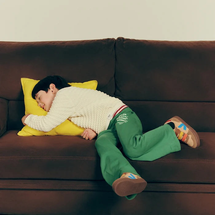 A child lying on a brown couch with a yellow pillow, wearing a cream knit sweater, green pants, and colorful sneakers.