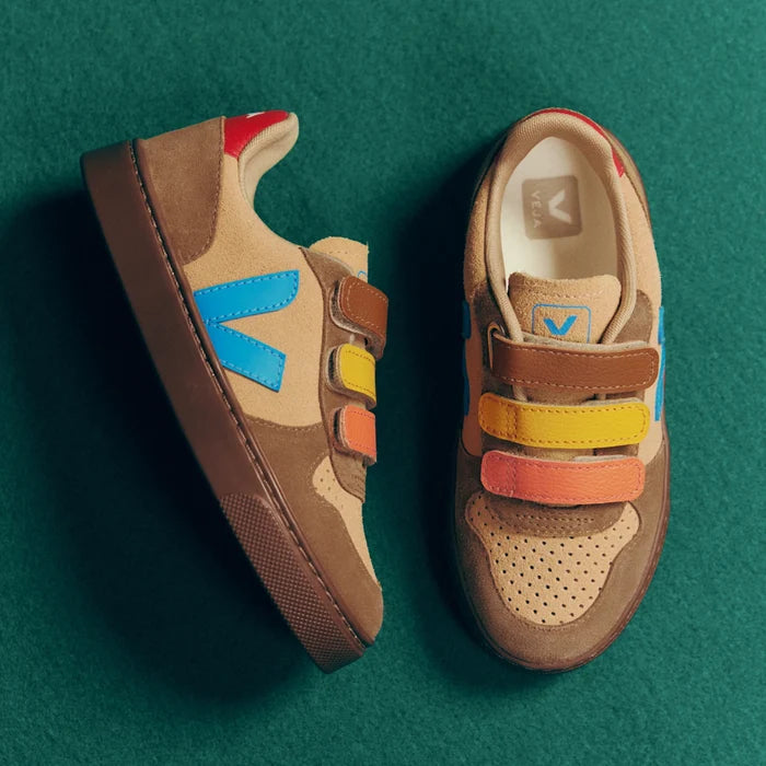 A pair of low-top sneakers shown from top and side views on a dark green textured background. The shoes feature a tan suede and mesh upper with brown overlays, colorful hook-and-loop straps in blue, yellow, orange, and red, and a brown sole.