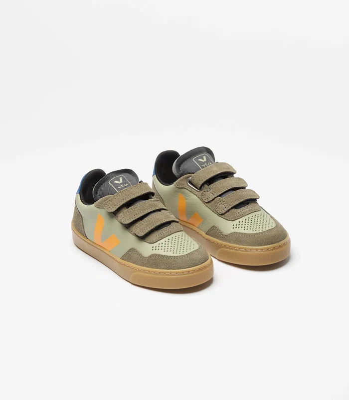 A pair of children's low-top sneakers in muted olive green and beige, featuring three wide hook-and-loop straps, a gum sole, and a small orange accent on the side, shown against a plain white background.