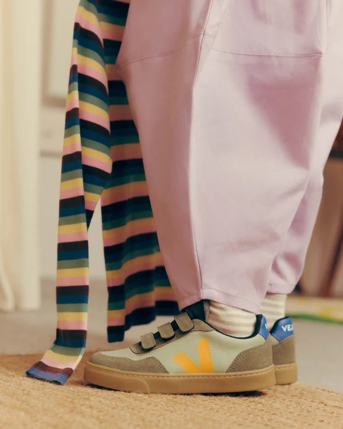 A close-up lifestyle shot of a person wearing light pink sweatpants and holding a pastel striped knit top, with their feet in green and orange Veja sneakers. The person is standing on a textured beige rug.