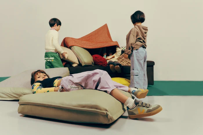 Four children play with oversized cushions and a small blanket fort in a minimalist indoor room with a white wall and green floor strip.