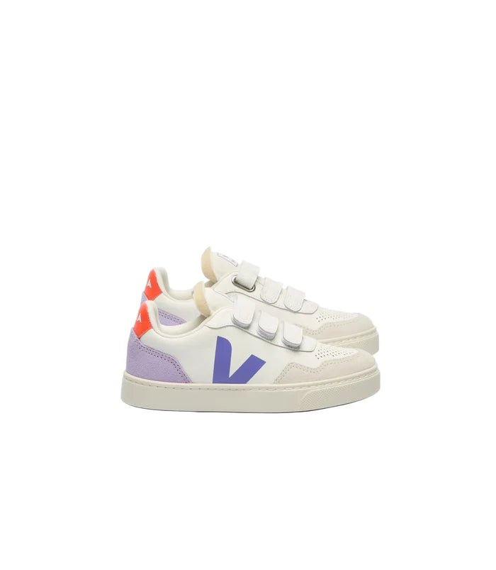 A pair of Veja Kids low-top sneakers in white leather, featuring a large purple 'V' logo on the side, double hook-and-loop straps, a purple heel tab, and a bright orange heel accent, set against a plain white background.