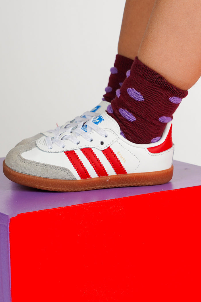 A white  sneaker with red stripes and a gray suede toe cap, worn with burgundy socks featuring purple polka dots, standing on a bright red block.