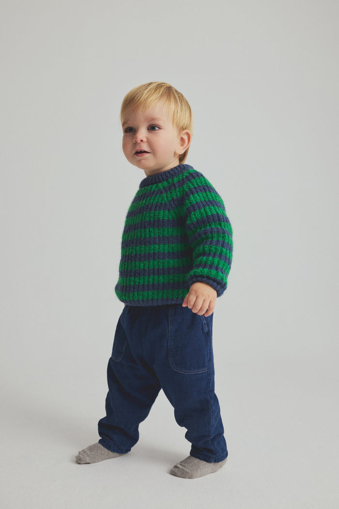 Child wearing a green and blue striped sweater and blue pants on a light gray background