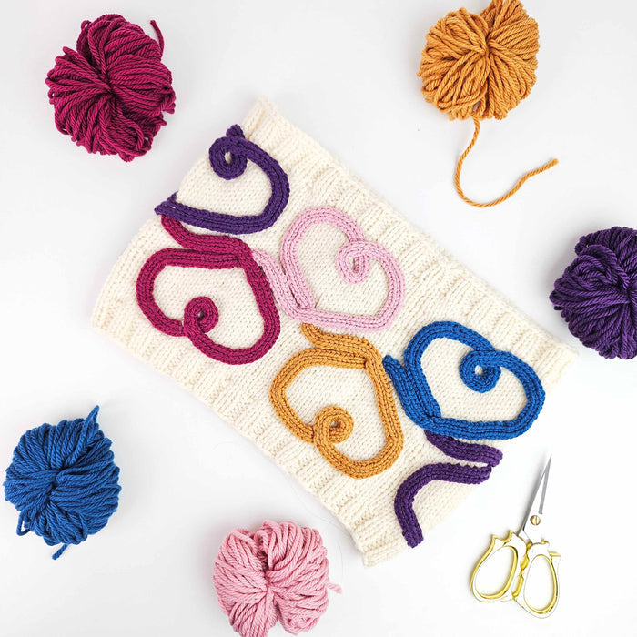 Flat lay of the finished Wandering Hearts Cowl in cream with swirling cord quill hearts in magenta, pink, gold, purple, and blue surrounded by coordinating yarn balls and gold scissors