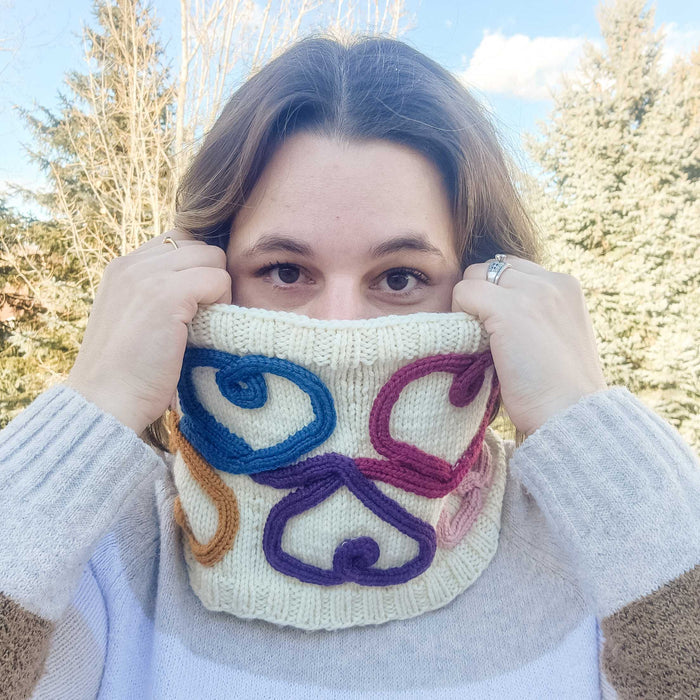 Woman wearing the Wandering Hearts Cowl pulled up over her nose showing the colorful cord quill heart design in blue, gold, magenta, pink, and purple on cream knitted fabric