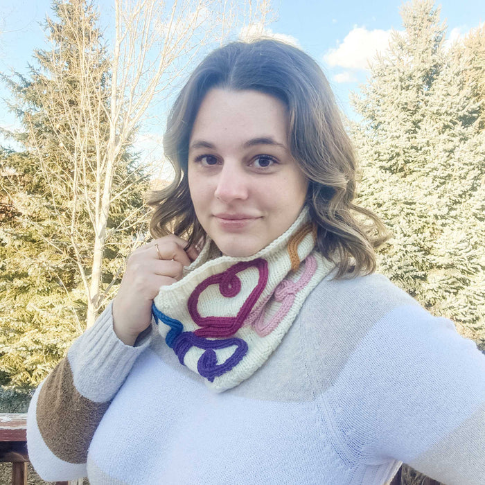Woman wearing the Wandering Hearts Cowl around her neck outdoors showing the cord quill heart design in magenta, pink, purple, and blue on cream knitted fabric