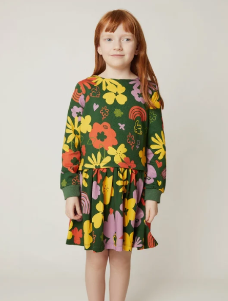 A young girl with red hair wearing a dark green long-sleeve dress with a colorful retro floral pattern, standing against a plain light beige background.