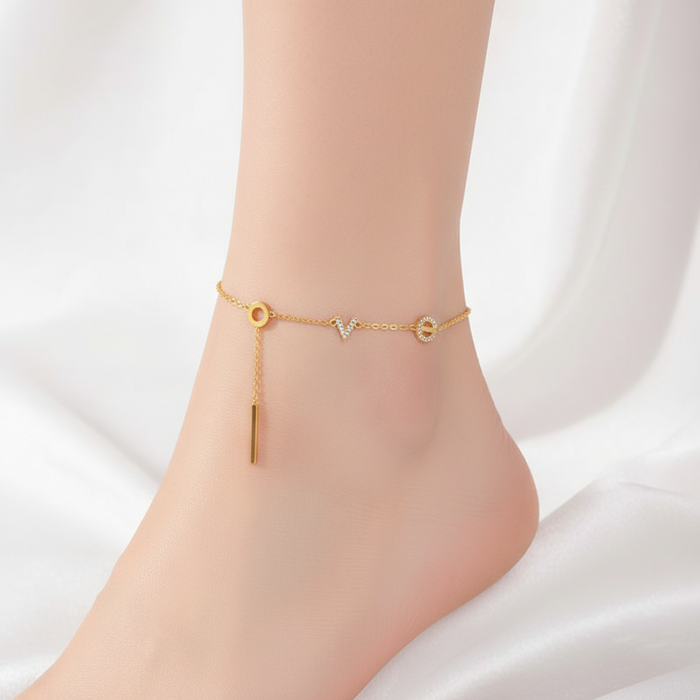Dainty gold chain anklet featuring crystalline letter charms spelling LOVE with a hanging bar pendant on a model's ankle