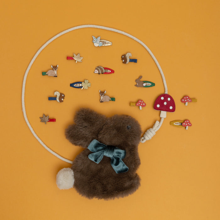 A plush brown bunny bag with a teal bow, attached to a white rope necklace with multiple woodland-themed charms, laid out on a solid orange background.
