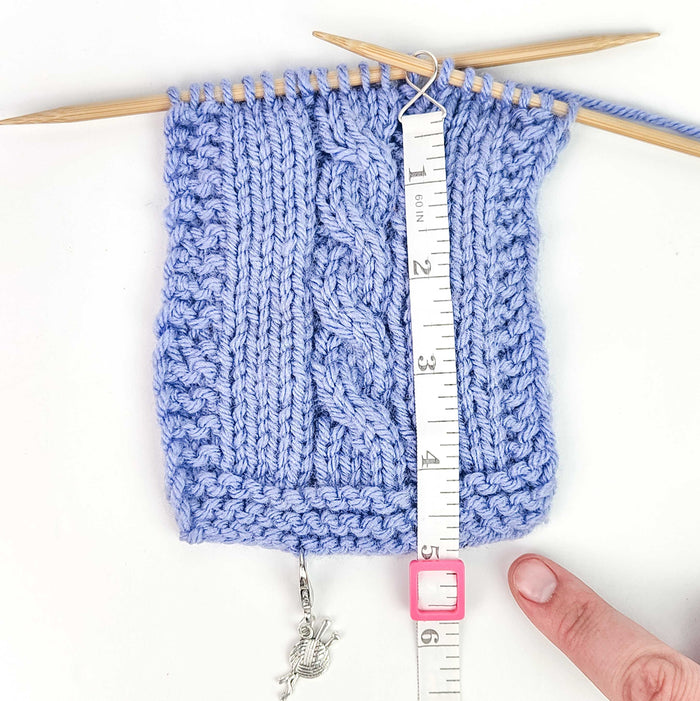 Work Until measuring tape hooked onto a blue cable knit swatch on bamboo needles showing the white retractable tape extended to measure rows with a pink square case and silver yarn ball charm