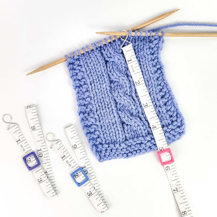 Work Until measuring tape in pink color hooked onto a blue stockinette knit swatch on bamboo needles showing the retractable white tape extended to measure rows