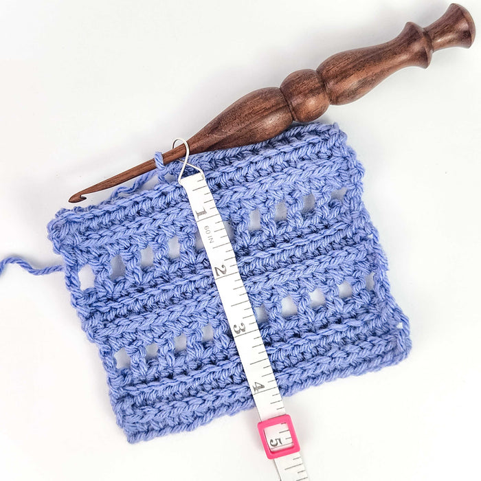 Work Until measuring tape in blue color hooked onto a blue stockinette knit swatch on bamboo needles showing the retractable white tape extended to measure rows