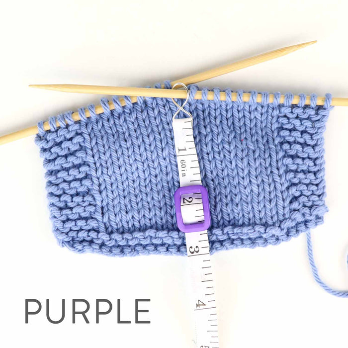 Work Until measuring tape in purple color hooked onto a blue knit swatch on bamboo needles showing the retractable white tape extended to measure rows