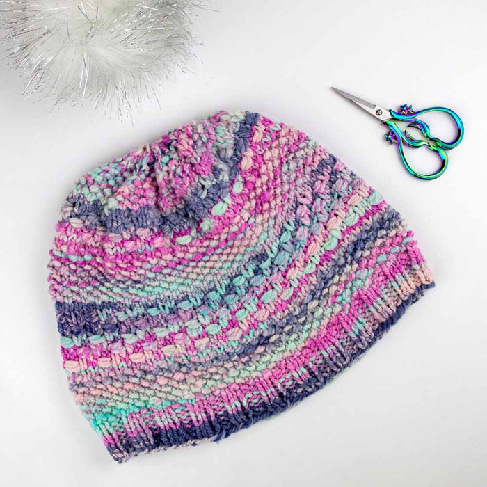 Multicolor knitted Wrapped Together beanie hat laid flat with rainbow scissors and faux fur pom pom