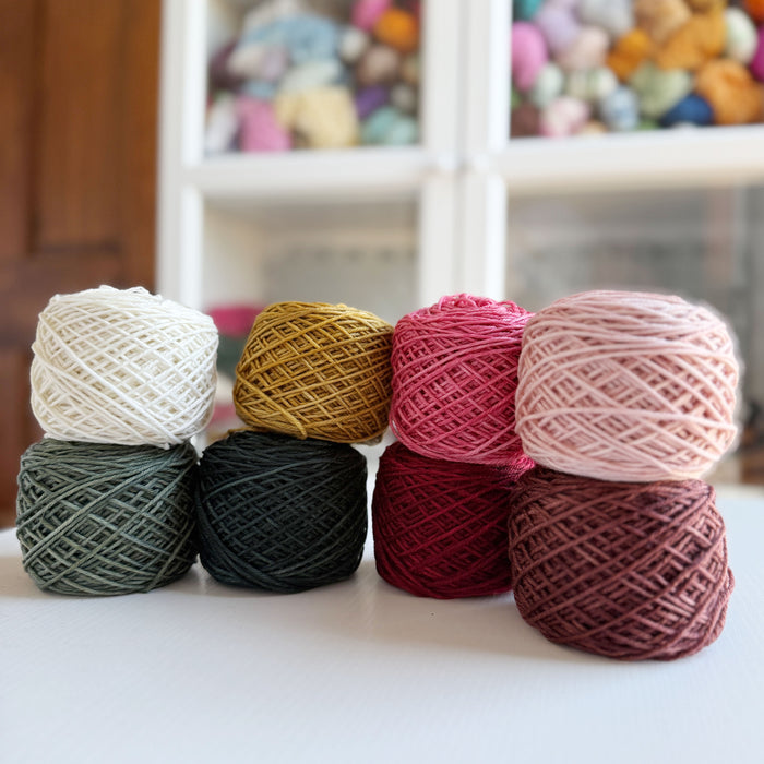 Eight yarn cakes of Yarnaceous Storybook DK Doodle yarn arranged in two rows of four on a white surface with a blurred yarn-filled glass hutch in the background showing center-pull cake format in Storybook colorways